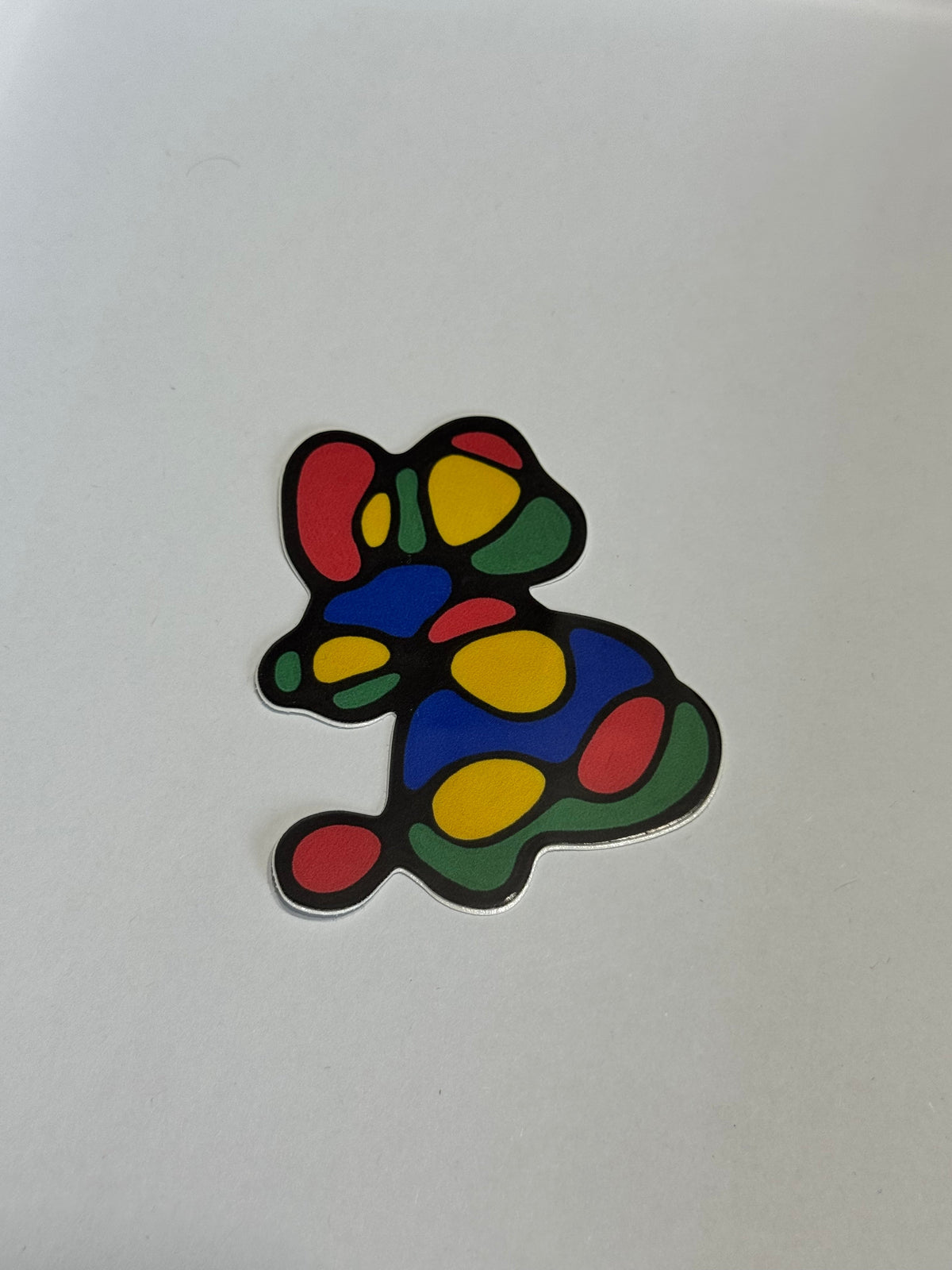 A small abstract mouse-shaped sticker or decal with a black outline and colorful filled spots in red, yellow, green, and blue, placed on a plain light background.